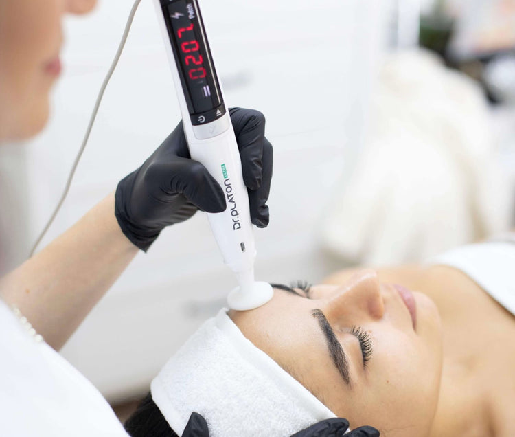 A close-up of a beauty practitioner wearing black gloves using a white and black LED facial treatment device on a client's forehead. The practitioner is wearing a white uniform, and the client is lying down with a white head towel.